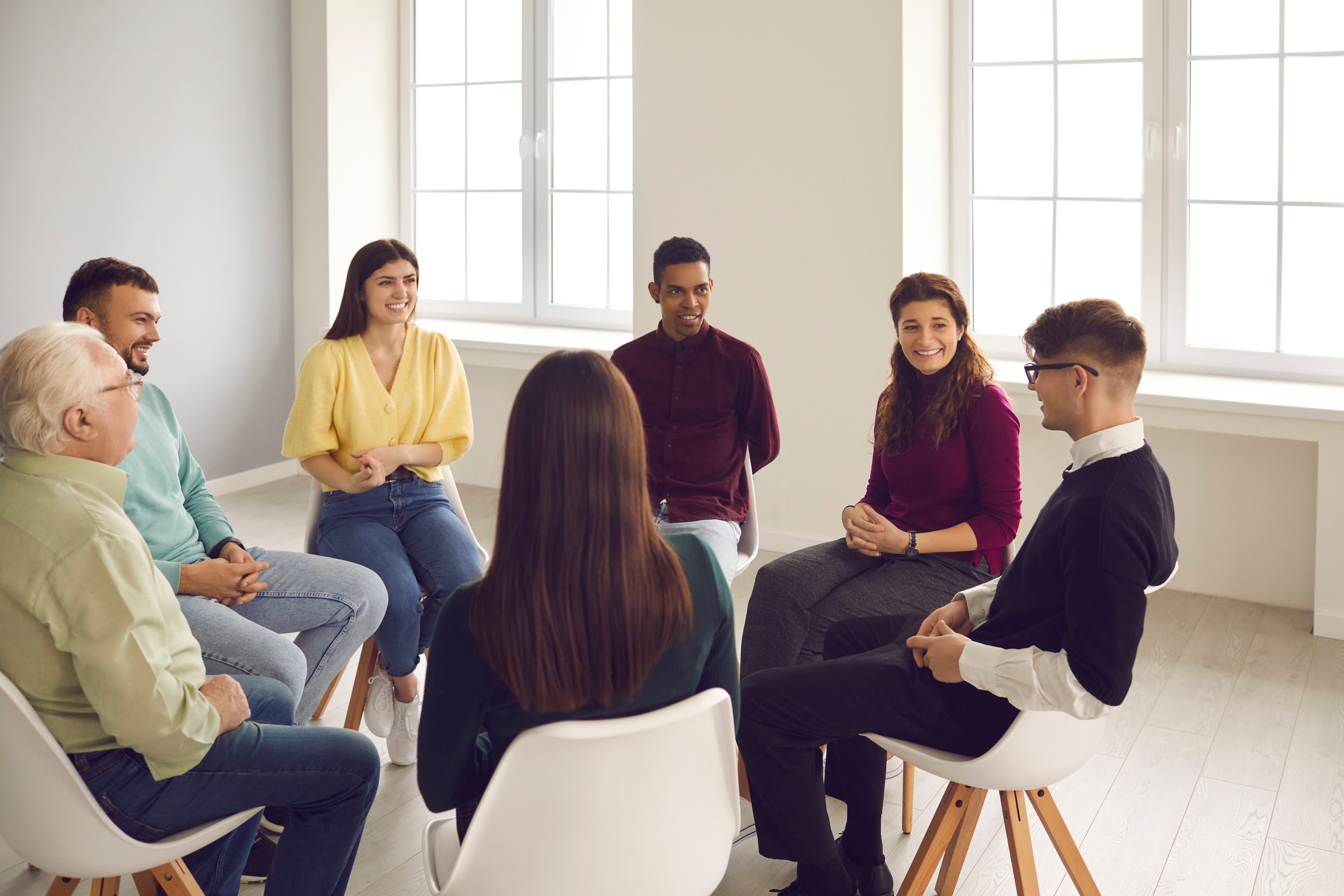 Getting psychological help in a friendly atmosphere. Happy smiling diverse people sitting in circle and listening to young man talking about his concerns and sharing opinion in a group therapy session