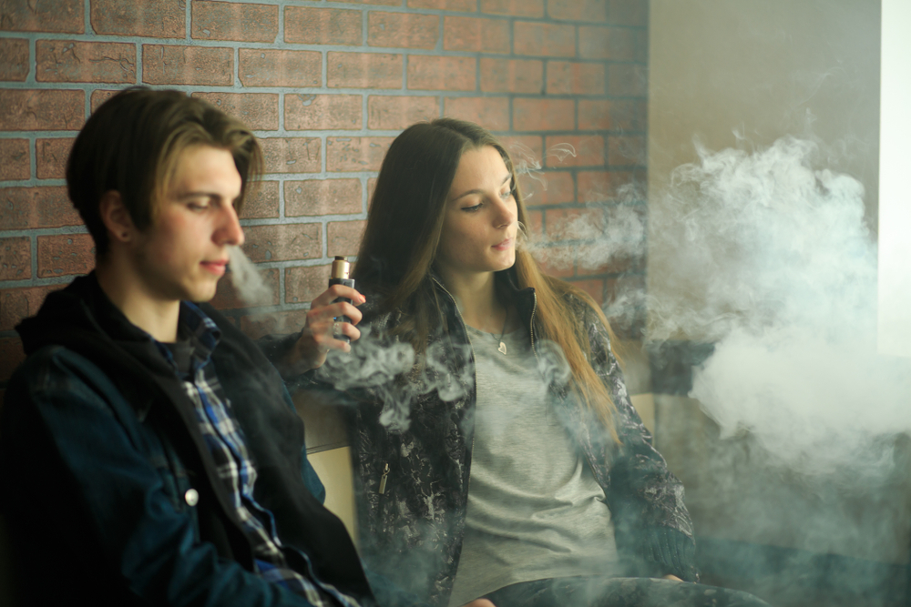 Vape teenagers. Young cute girl in sunglasses and young handsome guy smoke an electronic cigarettes in the vape bar. Bad habit that is harmful to health. Vaping activity.